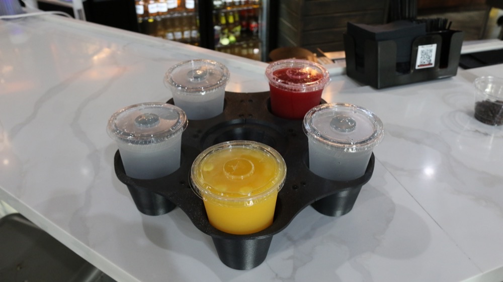 Top-down view of the GrippyTray on a marble bar: five molded cup wells around a central grip, four cups locked into the wells with lids on