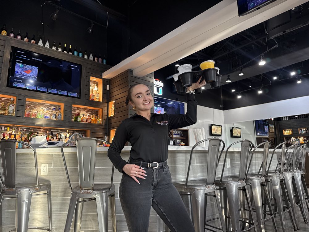 Server raising the loaded GrippyTray overhead with one hand at a bar, drinks fully secured in the wells
